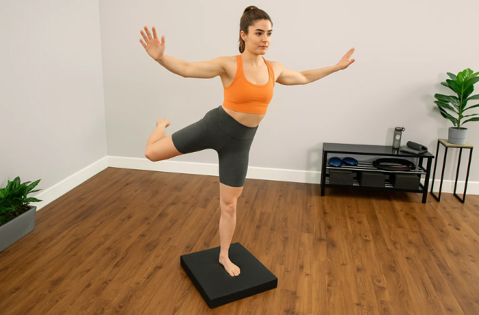 Woman performing dancer’s pose on a black foam pad in a fitness room