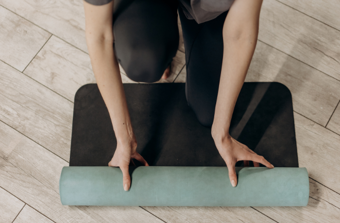 Person kneeling while unrolling a two-tone natural-rubber yoga mat (aqua surface, black base) on a parquet floor
