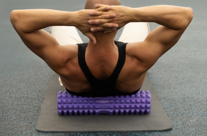 Man with muscular build rolling his upper back on a purple textured foam roller during a gym workout.