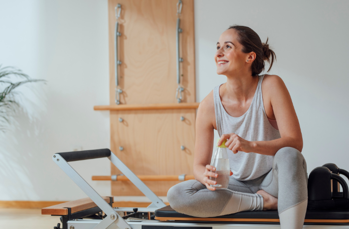 A woman smiling while sitting on a Pilates reformer machine, holding a water bottle, in a well-lit studio.